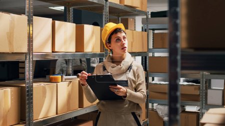 Photo for Young woman writing stock logistics in warehouse, looking at cardboard boxes on shelves. Female storage room employee using papers to check merchandise for industrial distribution. - Royalty Free Image
