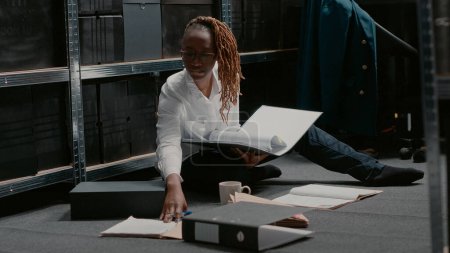 Photo for Focused law officer examining case files in folder, reading important criminal clues on police archive floor. Female inspector doing research to find suspect with surveillance photos. Handheld shot. - Royalty Free Image
