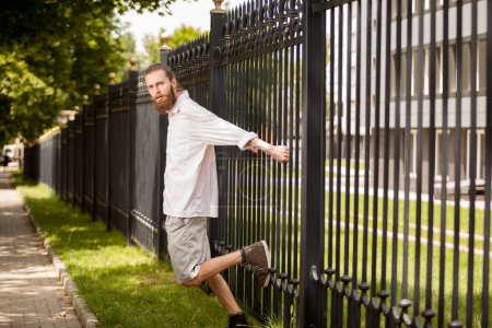 Photo for Bearded hipster posing next to a fence outside in the city. Style and diversity - Royalty Free Image