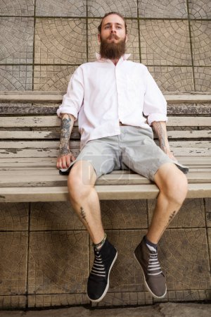 Téléchargez les photos : Hipster tatoué barbu couché sur un banc. Style et mode - en image libre de droit
