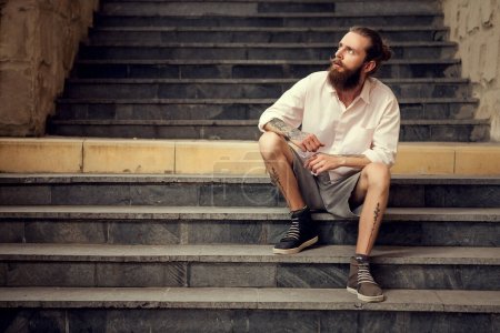 Foto de Beardead tatuado fresco buscando chico sentado en la calle en las escaleras mirando lejos de la cámara - Imagen libre de derechos
