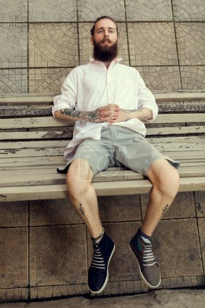 Téléchargez les photos : Cool gars à la recherche avec des tatouages et longue barbe assis sur la chaise et posant dans le style de la mode. Non-conformisme et hipster - en image libre de droit