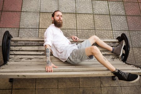 Foto de Un hipster tatuado acostado en un banco. Estilo y moda - Imagen libre de derechos