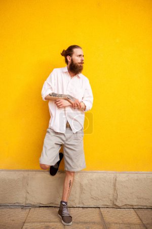 Téléchargez les photos : Tatoué homme attrayant barbu sur mur jaune posant en plein air - en image libre de droit