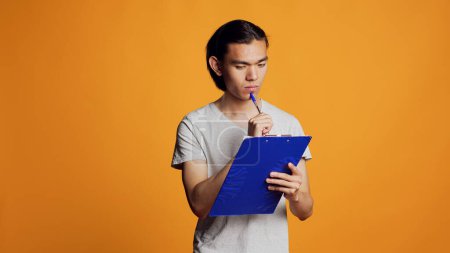 Photo for Confident young man writing and taking notes on files, using clipboard with papers to have informations. Carefree modern person holding pen to sign documents in studio, male model. - Royalty Free Image