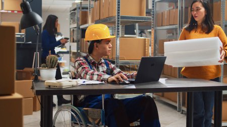 Téléchargez les photos : Young man in wheelchair looking at stock supplies, checking merchandise in boxes with female worker. Asian adult with impairment planning business development in storage room. - en image libre de droit
