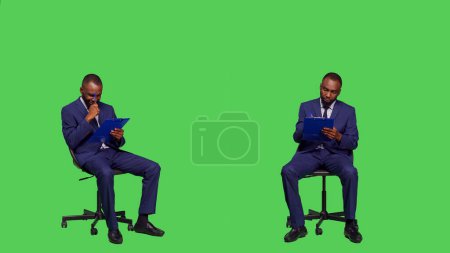 Téléchargez les photos : Un homme d'affaires afro-américain prend des notes sur des dossiers, réfléchit à de nouvelles idées d'entreprise sur fond d'écran vert. Homme entrepreneur écrivant des informations commerciales sur les papiers, assis sur la chaise. - en image libre de droit