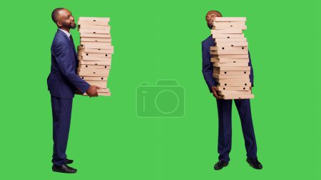Téléchargez les photos : Employé de bureau transportant une grosse pile de pizzas en studio, tenant un repas de restauration rapide sur fond vert. Startup homme d'affaires en costume ayant ordre à emporter sur la caméra. - en image libre de droit