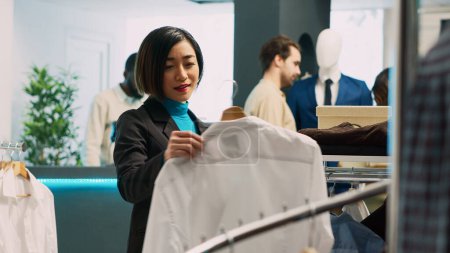 Téléchargez les photos : Client asiatique regardant chemise de vêtements formels dans le magasin de détail, en utilisant des marques à la mode pour augmenter la garde-robe. Femme adulte achetant des marchandises de boutique de mode, concept de petite entreprise. - en image libre de droit