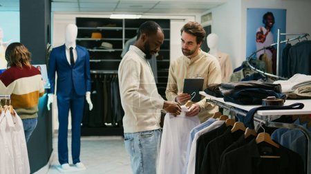 Photo for African american man checking merchandise on hangers, asking store assistant about new fashion collection. Young client talking to mall employee about clothes in retail shop boutique. - Royalty Free Image