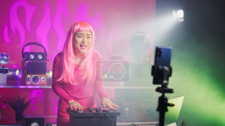 Foto de Cheerful artist playing techno song using mixer console, recording music session with mobile phone camera. Dj woman using electronics to do musical performance using audio equipment - Imagen libre de derechos