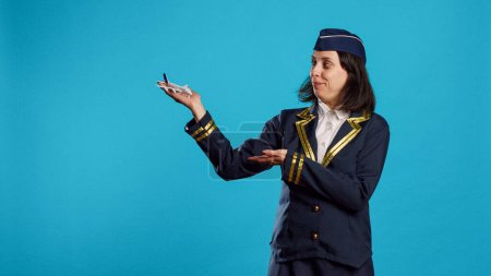 Photo for Smiling stewardess showing artificial plane toy, feeling confident about aviation profession. Young person working as air hostess in uniform showing fake miniature plane in studio. - Royalty Free Image