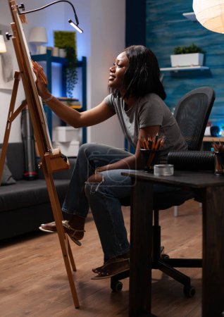 Foto de Dibujo profesional femenino haciendo obras de arte a mano con lápices, sentado al lado de herramientas creativas de trabajo. Mujer afroamericana con habilidades artísticas creando ilustración sobre lienzo en blanco. - Imagen libre de derechos