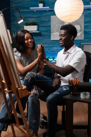 Photo for Partners with artistic abilities giving high five as they finish drawing with colored pencils on canvas easel in art studio. African american sketching teacher congratulating student for masterpiece. - Royalty Free Image