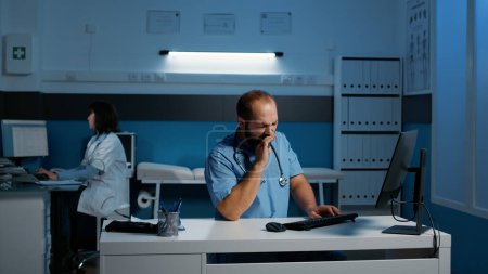 Photo for Tired exhausted physician assistant typing medical expertise on computer while working over hours at patient medication treatment. Clinical staff checking report in hospital office - Royalty Free Image