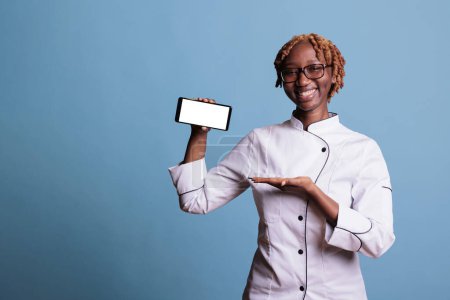 Téléchargez les photos : Femme afro-américaine portant un uniforme de cuisine montrant un téléphone portable avec écran blanc en studio. Cuisinier tenant appareil mobile regardant verticalement la caméra sur fond bleu. - en image libre de droit