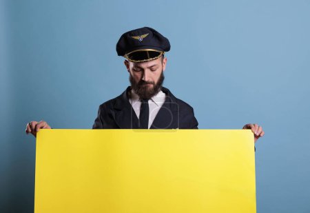 Téléchargez les photos : Aviateur d'avion tenant la bannière publicitaire vide jaune avec l'espace de copie, pilote regardant la plaque vierge dans les mains. Capitaine d'avion en uniforme d'aviation debout avec des annonces promotionnelles maquette - en image libre de droit