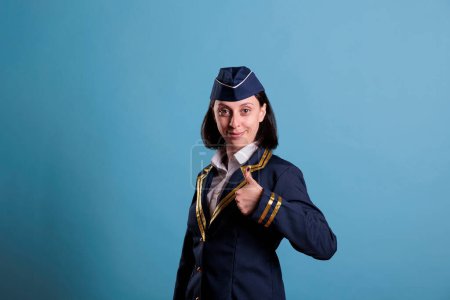 Photo for Portrait of smiling flight attendant making thumbs up gesture while standing in studio with blue background. air hostess wearing professional aviation uniform doing approval sign, medium shot - Royalty Free Image