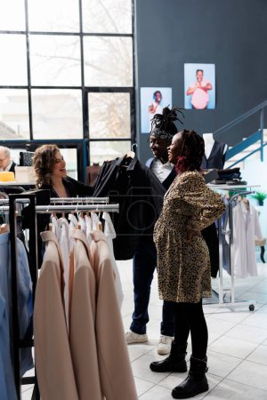 Photo for Employee helping diverse couple choosing right shirt for formal wear in shopping centre. Customers discussing clothes material with worker before buying merchandise in modern boutique - Royalty Free Image