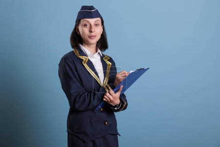 Photo for Smiling stewardess holding clipboard, writing, looking at camera. Young attractive flight attendant in aviation uniform filling form in airport, studio medium shot on blue background - Royalty Free Image