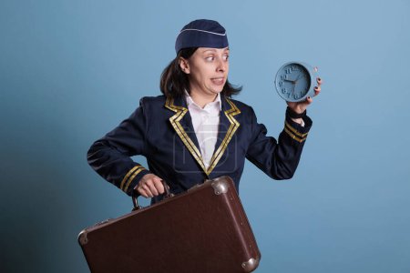 Photo for Flight attendant running late at airport, holding retro alarm clock, carrying suitcase. Airplane stewardess overslept, looking at time, air hostess hurry up at work with luggage - Royalty Free Image
