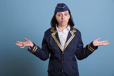 Photo for Portrait of confused flight attendant showing unsure gesture looking into camera. Pensive stewardess shrugging shoulders, making doubtful expressionin studio with blue background - Royalty Free Image