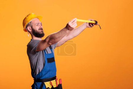 Photo for Confident construction worker using tape measure to take measurements and work on refurbishment project. Young male builder holding tapeline and measuring for renovation, wearing tools belt. - Royalty Free Image