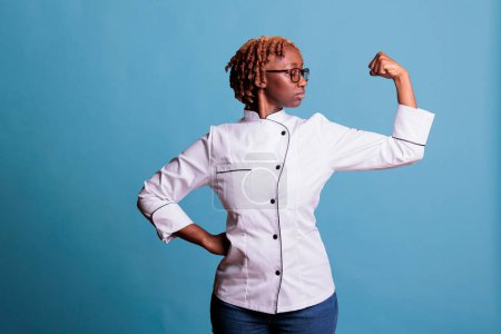 Téléchargez les photos : Femme cuisinière professionnelle bras fléchissant avec pose de puissance et de force en studio tourné. Chef afro-américain portant uniforme de cuisine regardant muscles du bras gauche sur fond bleu. - en image libre de droit