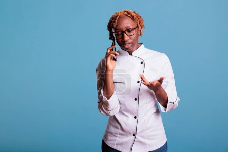 Photo for African american woman in chefs uniform having serious conversation with caterer for restaurant kitchen. Professional cook in studio shot against blue background. - Royalty Free Image