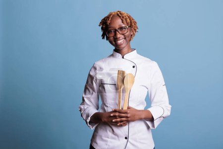 Téléchargez les photos : Chef afro-américaine optimiste tenant des ustensiles de cuisine en studio tourné sur fond bleu. Cuisinière professionnelle sur le lieu de travail posant souriant pour la caméra. - en image libre de droit