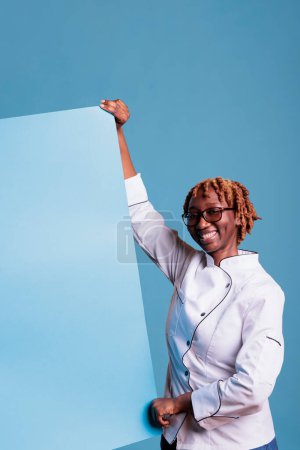 Photo for Young afro chef showing an ad. Happy kitchen helper, uniformed african american, holding a signboard, notice, publicity placard. Female member of restaurant doing marketing. - Royalty Free Image