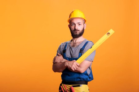 Photo for Construction worker holding water level ruler in studio, posing with arms crossed. Young repairman preparing to do building measurements using professional leveler tool, confident carpenter. - Royalty Free Image
