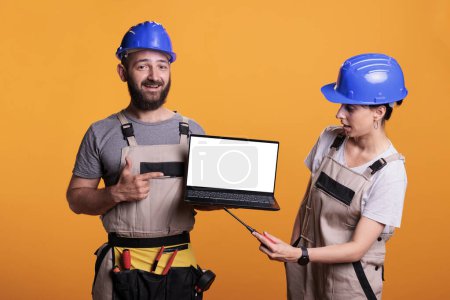 Cheerful renovators pointing at white laptop screen, showing isolated blank display on wireless pc. Team of builders holding computer with empty copyspace template, posing in studio.