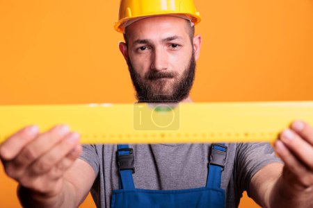 Photo for Confident male carpenter looking at construction leveler before working on refurbishment project, studio shot. Handyman builder carrying water level tool or ruler to work on renovation. - Royalty Free Image
