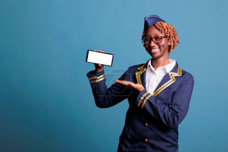 Photo for African american stewardess holding smartphone with white screen in a studio showing advertising space. Female flight attendant in uniform with cell phone with blank copy space. - Royalty Free Image