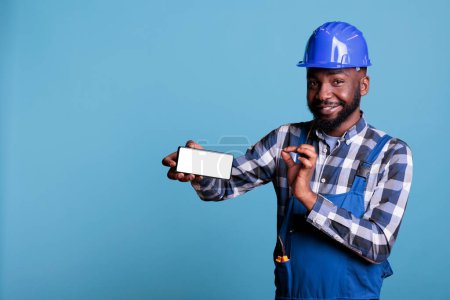 Téléchargez les photos : Homme en uniforme de travail tenant smartphone avec écran publicitaire vierge en studio tourné. Travailleur de la construction afro-américain pointant vers écran d'affichage vide de téléphone portable sur fond bleu. - en image libre de droit