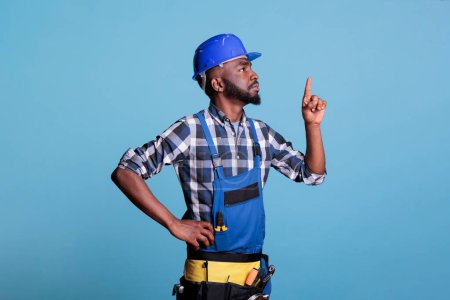 Téléchargez les photos : Constructeur afro-américain pointant vers le haut en studio tourné sur fond bleu, portant un uniforme de travail et un casque tout en effectuant une rénovation intérieure. Concept de rénovation et d'amélioration. - en image libre de droit