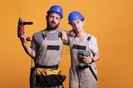 Photo for Team of professional engineers holding power drill guns in studio, posing with electric tools over background. Man and woman using drilling gun and wearing overalls with hardhat, tools belt. - Royalty Free Image
