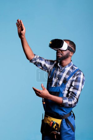Téléchargez les photos : Travailleur de la construction portant des lunettes de réalité virtuelle sur fond bleu. Constructeur afro-américain bénéficiant de divertissement 3D offert par la technologie moderne. - en image libre de droit