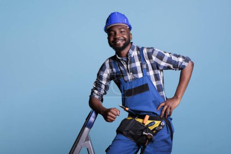 Photo for Optimistic builder looking at camera with big smile while resting after finishing work in studio shot against blue background. Contractor using tool belt needed for renovation construction tasks. - Royalty Free Image