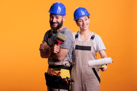 Photo for Team of contractors with paintbrush and roller in studio, working on painting job with tools. Construction workers experts wearing overalls and posing with brushes, doing renovation project work. - Royalty Free Image