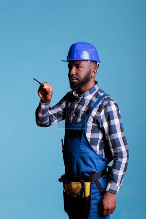 Foto de Retrato de un profesional de la construcción de un destornillador lateral sobre fondo azul. Constructor afroamericano concentrado que usa cinturón de herramientas y uniforme de trabajo. - Imagen libre de derechos