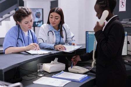 Photo for Hospital reception worker talking at landline phone being responsible for scheduling appointments and managing patient records. Nurse and doctor looking at papers with medical expertise in lobby - Royalty Free Image