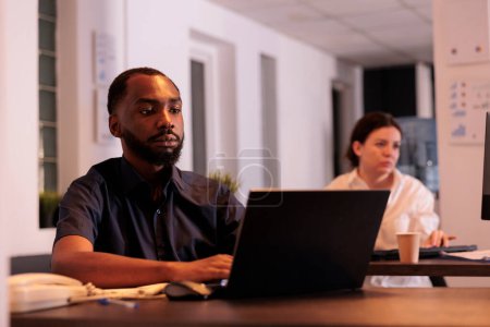 Téléchargez les photos : Employé utilisant un ordinateur portable d'entreprise dans un bureau de coworking, travailleur assis au bureau du lieu de travail. Homme d'affaires afro-américain travaillant sur le rapport de projet de démarrage sur l'ordinateur la nuit - en image libre de droit