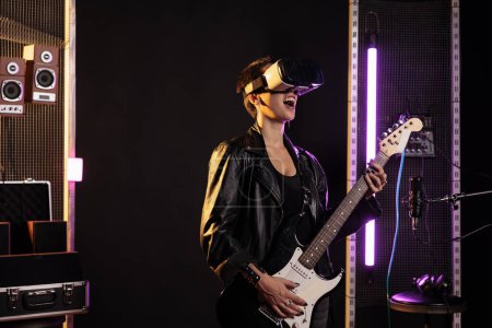 Photo for Rockstar with vr goggles enjoying rock concert simulation while playing heavy metal song at electric guitar in music studio. Woman musician performing grunge album using electricinstrument - Royalty Free Image