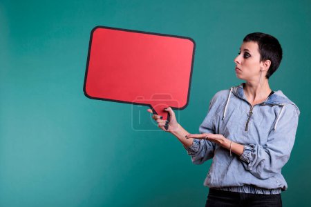 Photo for Attractive woman looking at camera while pointing at empty red speech bubble standing in studio over isolated background. Cheerful confident female with blank sign for communication on board - Royalty Free Image