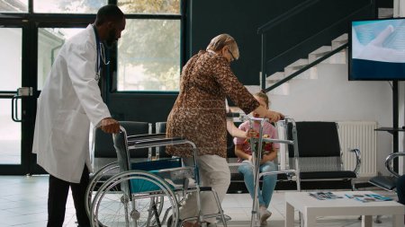 Téléchargez les photos : Femme âgée en fauteuil roulant parlant à une petite fille dans la salle d'attente, famille venant rendre visite à une patiente âgée ayant une déficience. Utilisateur de fauteuil roulant essayant de marcher avec cadre de marche dans le hall de l'hôpital. - en image libre de droit
