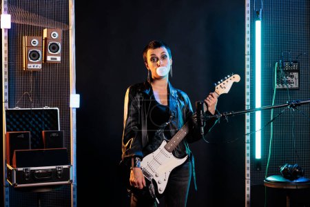 Photo for Brunette rockstar woman with short hair and grunge style blowing bubble gum while playing electric guitar preparing for rock concert. Guitarist using electric instrument to performing heavy music - Royalty Free Image