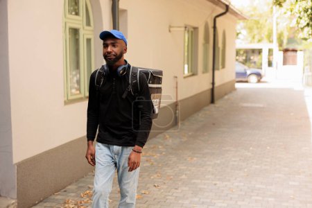 Photo for Restaurant takeaway meal african american courier walking in street, standing outdoors, front view. Food delivery by foot, deliveryman going with customer order in thermal backpack - Royalty Free Image