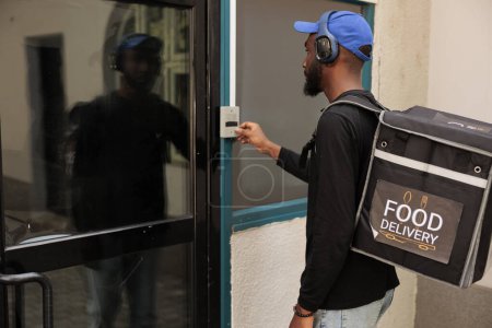 Photo for Business lunch delivery service courier ringing office building doorbell, waiting for client outdoors. Young african american man with backpack delivering takeaway restaurant meal - Royalty Free Image